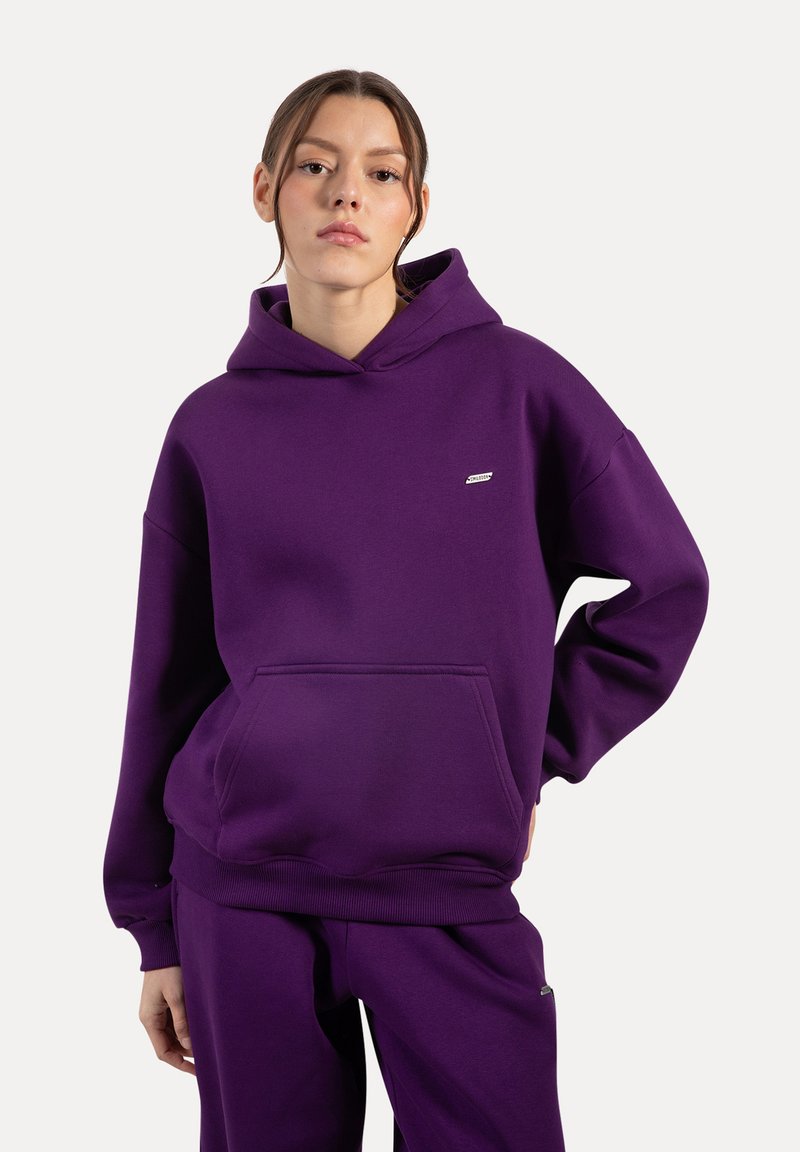 Sudadera morada de tejido suave con un bolsillo tipo canguro. Presenta un dobladillo y puños acanalados, y un pequeño logo sobre el pecho izquierdo.
