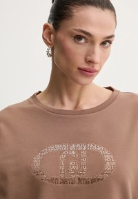 Felpa marrone con scollo rotondo, caratterizzata da un logo decorativo composto da piccoli rivetti metallici sulla parte anteriore. Texture liscia e vestibilità rilassata.