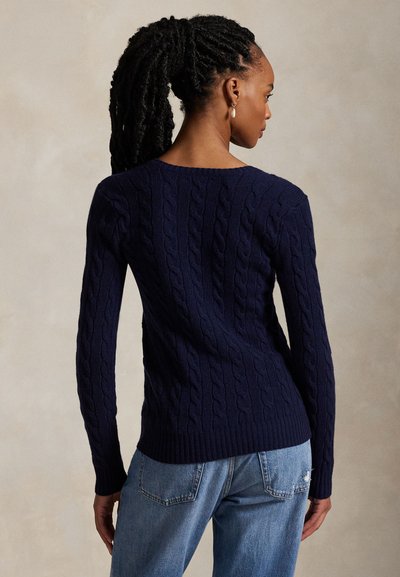 Woman with braided hair wearing navy cable-knit sweater and light blue jeans, facing away against neutral background.