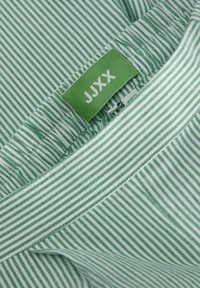 Zelenobílá pruhovaná látka s štítkem "JJXX." Materiál je lehký a má hladkou texturu.