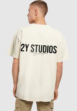 Vaalean kermaisuinen oversized t-paita, jossa on selässä suuret mustat tekstit "2Y STUDIOS" ja "VAIN YHTEISÖN JÄSENILLE". Lyhyet hihat.