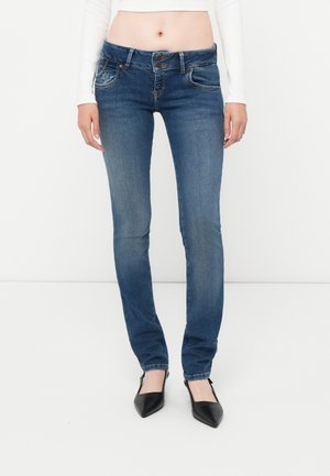 Woman wearing mid-rise blue skinny jeans with double button closure and black pointed-toe heels, standing against white background.