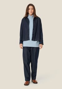 Denimjakke over lys blå ribbet sweater; løstsiddende mørke denimbukser; brune loafers; flade knapper på jakken og ribbet struktur i sweateren.