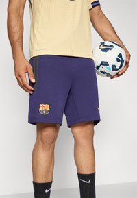 Nike Barcelona rövidnadrág, lila szövetből, oldalán cipzárakkal és csapatlogóval. Egy fehér focilabdával kísérve, kék részletekkel.