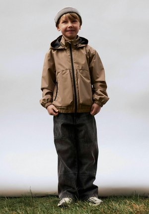 Nuori poika, joka on pukeutunut beigeen takkiin, raidalliseen pipoon, mustiin leveisiin housuihin ja valkoisiin lenkkareihin, seisoo nurmikolla vaaleaa taustaa vasten.