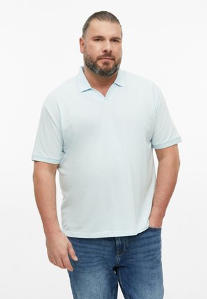 Brodaty mężczyzna z gładko zaczesanymi do tyłu włosami, ubrany w jasnoniebieską koszulkę polo z krótkim rękawem i niebieskie dżinsy, stoi na białym tle.