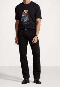 Muž oblečený v černých džínách, černém pásku, černých kožených botách, černých hodinkách a černém tričku s grafikou medvěda držícího dárek.