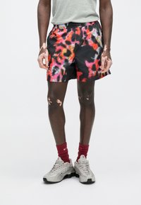 Μαύρο σορτς με έντονο κόκκινο, ροζ και πορτοκαλί αφηρημένο μοτίβο, ελαστική μέση, φορεμένο με κόκκινες κάλτσες και γκρι αθλητικά παπούτσια.