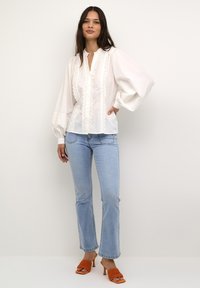 Weiße Bluse mit floralem Spitzenbesatz und gepufften, gefältelten Ärmeln, kombiniert mit hellblauen Schlagjeans und orangefarbenen High-Heels.