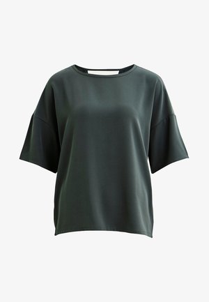 Mørkegrøn kortærmet bluse og hvide tætsiddende bukser, parret med sorte højhælede sko. Blusen har en rund halsudskæring og en glat tekstur.