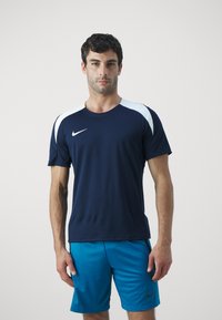 Marine atletisk t-skjorte med hvite skulderdetaljer, korte ermer og Nike-logo. Kombinert med blåstripete shorts. Pustende tekstur.
