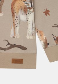 Felpa in tessuto beige con illustrazioni di un lynx e rami. Polsini e orlo in beige uniforme; etichetta in pelle con il marchio "walkiddy".