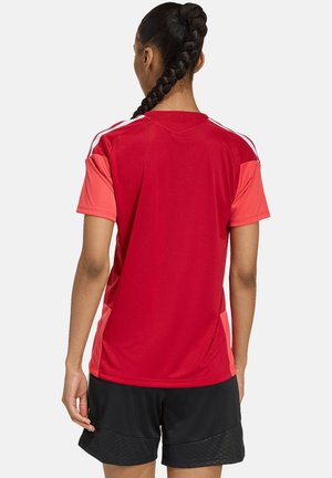 Osoba z zaplecionymi włosami, ubrana w czerwoną sportową koszulkę z jasnoczerwonymi rękawami i czarne spodenki, stoi tyłem na jednolitym tle.