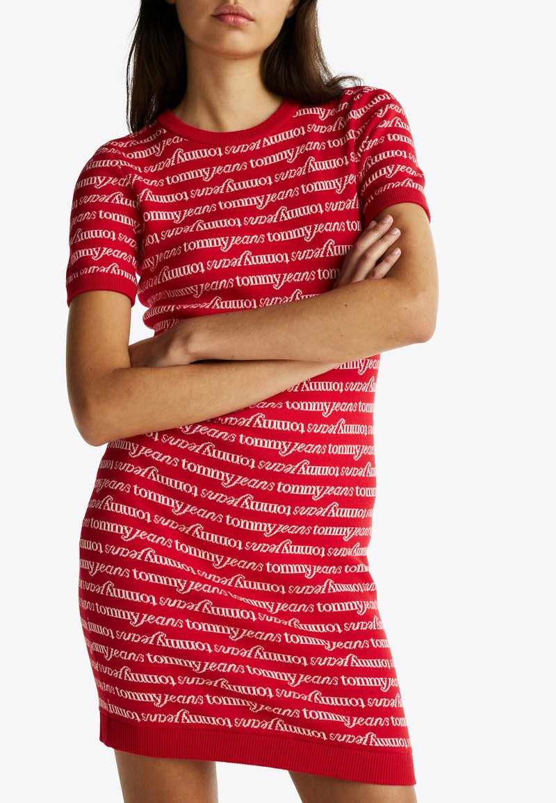 Rotes, figurbetontes Kleid mit kurzen Ärmeln, das ein wiederholendes weißes "Tommy Jeans"-Logomuster zeigt. Strukturiertes Gewebe mit geripptem Saum.