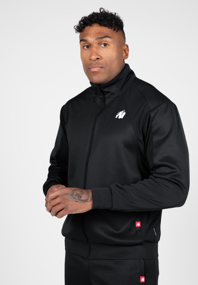 Zwarte sportjack met hoge kraag, ritsluiting, gestructureerde panelen en een wit logo. Inclusief rode accentlabels.