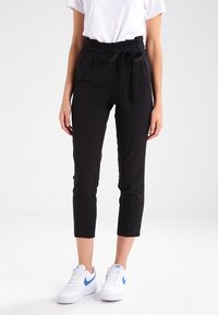 Svarta cropped byxor med hög midja, tygbälte och sidofickor, parat med en vit t-shirt och vita sneakers med blå detaljer.