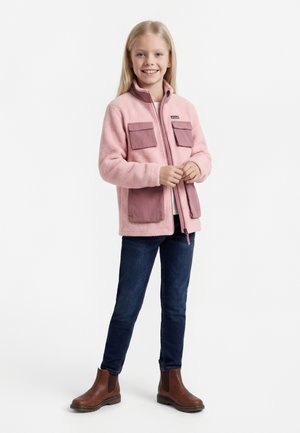 Jente iført en rosa fleecejakke med lommer, blå jeans og brune støvler, står og smiler mot en hvit bakgrunn.