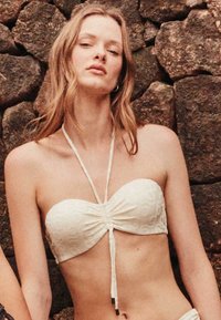 Witte bandeau bikini top met ruches in het midden, verstelbare banden en een gestructureerde stof. Draagt door een model tegen een stenen achtergrond.