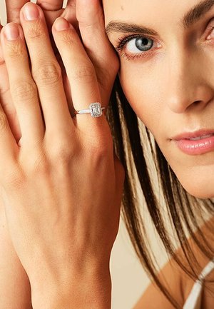 Eleganter Silberring mit einem smaragdgeschliffenen Zentrumstein, umgeben von kleineren Diamanten. Getragen an einer Hand mit natürlichen Nägeln. Weiche Hauttextur.