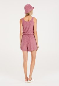 Rompers texturé rose avec une taille cintrée et un chapeau seau assorti. Présente un design sans manches et des jambes courtes. Des sandales blanches complètent le look.