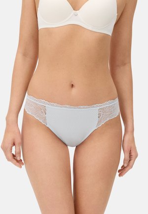 Mujer con sujetador blanco y bragas blancas con ribete de encaje, de pie con los brazos relajados a los lados contra un fondo liso.