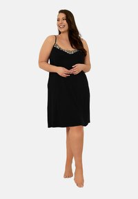 Uśmiechnięta kobieta z długimi brązowymi włosami, ubrana w czarną sukienkę bez rękawów z lamówką w leoparda, stojąca na boso na neutralnym tle.