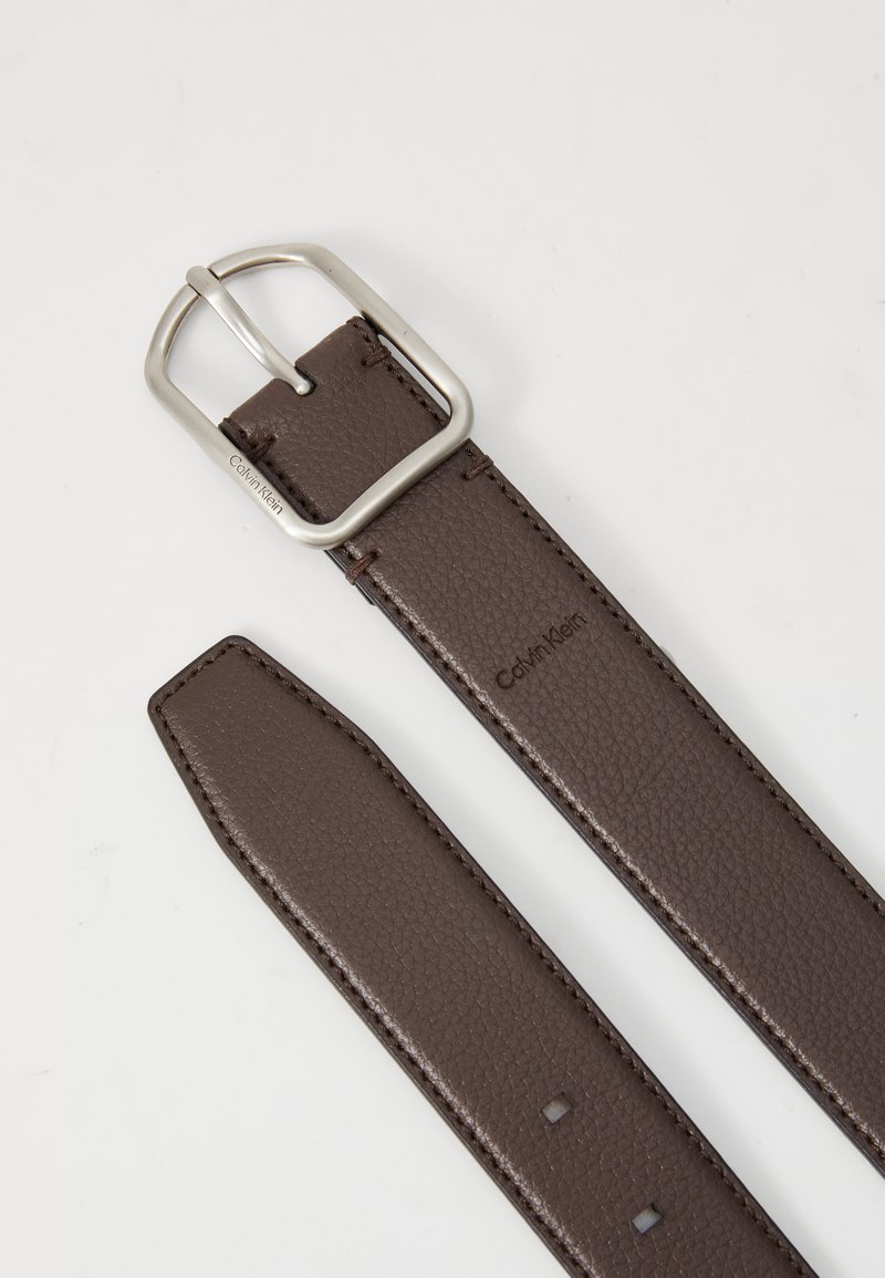 Ceinture en cuir brun avec une surface texturée, dotée d'une boucle en métal argenté et d'un petit logo gravé. Découpe droite sans motifs.