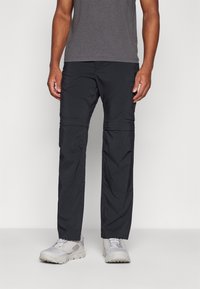 Pantalons de randonnée noirs à zip, fabriqués dans un tissu léger et résistant à l'eau, avec une texture lisse et des poches latérales ; associés à des chaussures de randonnée grises.