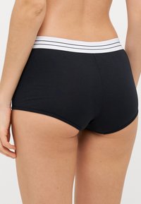Svarta bomullsliknande boyshorts med ett vitt elastiskt midjeband som har två tunna svarta ränder. Tyget har en slät textur.