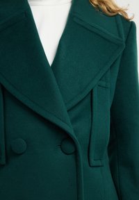 Abrigo de lana en color verde azulado, de doble botonadura, con grandes solapas, bolsillos decorativos y botones redondos. La tela texturizada completa el diseño estructurado.