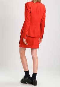 Röd kavaj och matchande minikjol. Kavajen har en skräddarsydd passform och knappar, medan kjolen är rak med en subtil textur. Svarta skor fulländar looken.