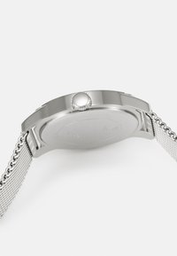 Silberne Edelstahl-Uhr mit einem Netzband, ausgestattet mit einem runden Gehäuse und einer strukturierten Krone. Der Gehäuseboden ist glatt und unbeschriftet.