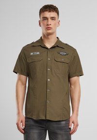 Kortermet olivengrønn skjorte med to brystlommer, knappelukking foran, skulderklaffer og broderte merkeaksenter.