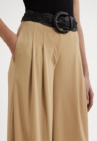 Czarny, pleciony skórzany pasek z dużą, okrągłą sprzączką, noszony na beżowych plisowanych spodniach z kieszeniami po bokach.