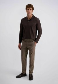 Man staat met één hand in zijn zak, gekleed in een donkerbruine poloshirt met lange mouwen, lichtbruine broek en donkere nette schoenen, tegen een eenvoudige achtergrond.