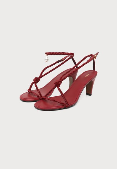 Sandales rouges à talons hauts avec des brides torsadées en corde et un petit détail en perle sur la bride de cheville, affichées sur fond blanc.
