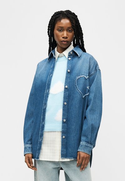 Femme aux longues tresses portant une veste en denim bleue avec un cœur effiloché sur la poitrine, par-dessus un pull bleu ciel avec des nuages blancs.