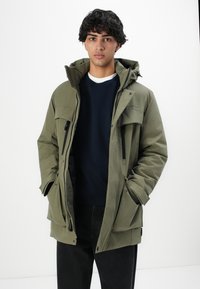 Junger Mann, der eine offene olivgrüne Parka mit mehreren Reißverschlusstaschen, schwarze Hosen und einen marineblauen Pullover vor einem weißen Hintergrund trägt.