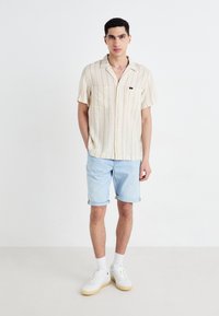 Short-sleeved, light beige shirt with vertical stripes, two front pockets. Paired with light blue denim shorts and white sneakers.