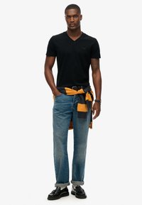 Sort v-hals t-shirt, lyseblå jeans og sorte loafers. Gul og gråternet sweatshirt bundet om taljen. Enkelt, afslappet outfit.