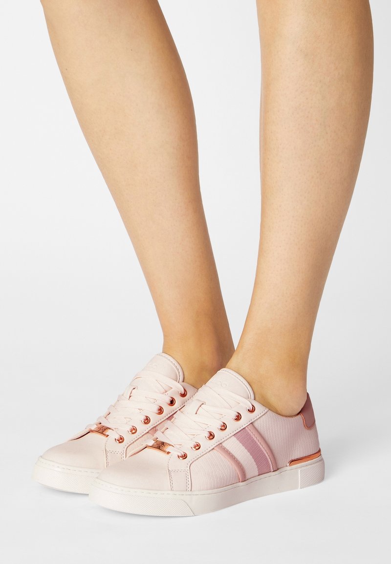 Baskets rose clair à finition lisse, avec des bandes roses, des œillets en or rose, et une tige en tissu texturé. Semelle en caoutchouc blanc.