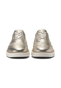 Un paio di brogue in pelle dorata metallizzata con lacci, dotate di suola bianca a righe, mostrate frontalmente su uno sfondo bianco.