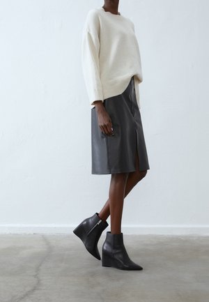 Pull en tricot crème, jupe en cuir gris avec une fente, et bottes à talons compensés noires. Textures lisses et détails de design taillés visibles.