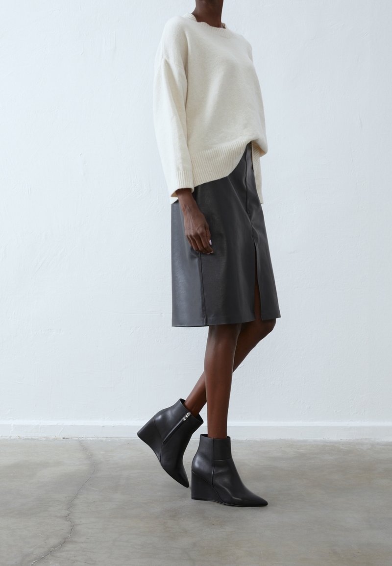 Pull en tricot crème, jupe en cuir gris avec une fente, et bottes à talons compensés noires. Textures lisses et détails de design taillés visibles.