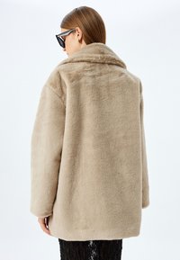 Abrigo de pelo sintético beige con un amplio cuello, ajuste oversize y textura suave. Ligeramente más largo en la parte de atrás, con detalles de costura visibles.