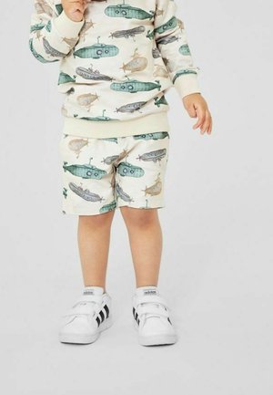 Barn iført en beige sweatshirt og shorts med ubådmønstre, med grønne, blå og brune accenter, parret med hvide sneakers.