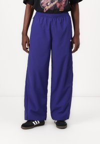 Pantalones de pierna ancha morados con cintura elástica, hechos de un material ligero, con bolsillos laterales y un acento de logo en el lado izquierdo.