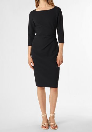 Kobieta ubrana w czarną, dopasowaną sukienkę do kolan z rękawami do połowy przedramienia oraz beżowe sandały na wysokim obcasie z paskami.