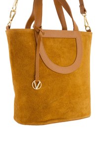 Tan suède draagtas met leren banden, voorzien van een afgeronde uitsnijding en een goudkleurig logo-embleem dat aan een band is bevestigd.