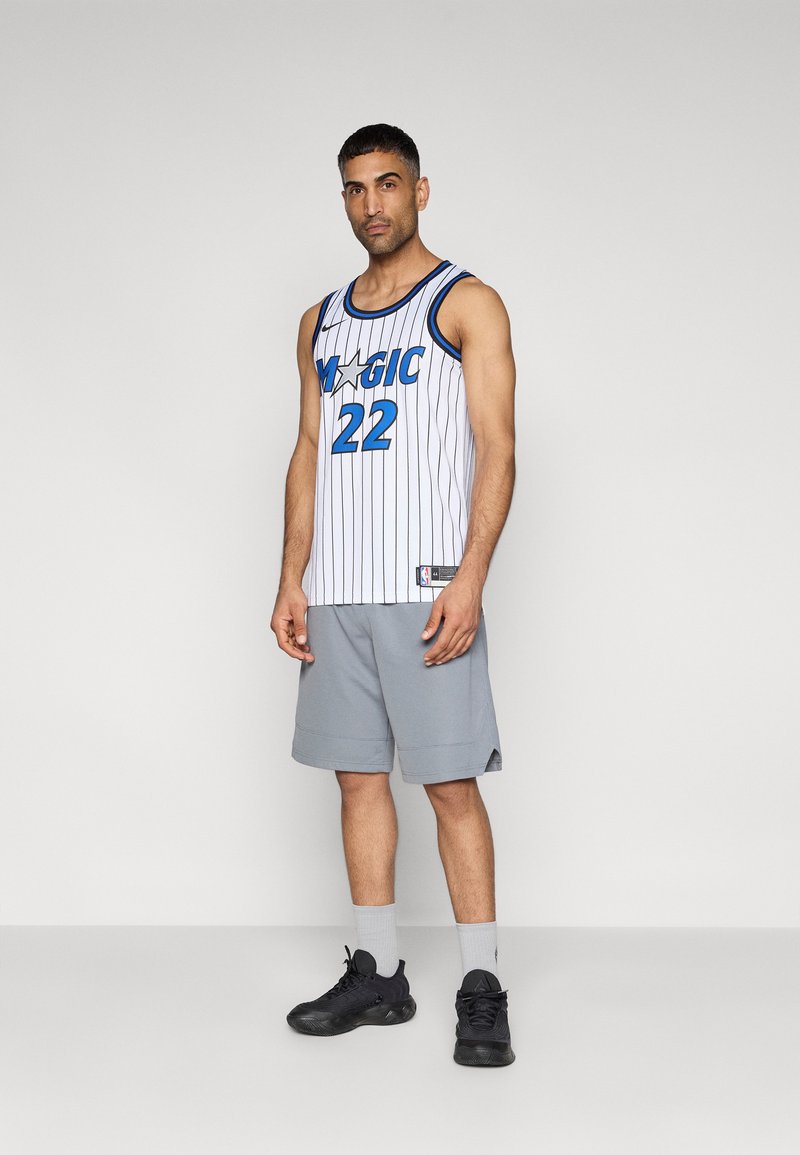 Herren-Basketballtrikot in Weiß mit blauen Nadelstreifen, mit der Aufschrift "MAGIC" und der Nummer 22. Kombiniert mit grauen Shorts und schwarzen Sportschuhen.